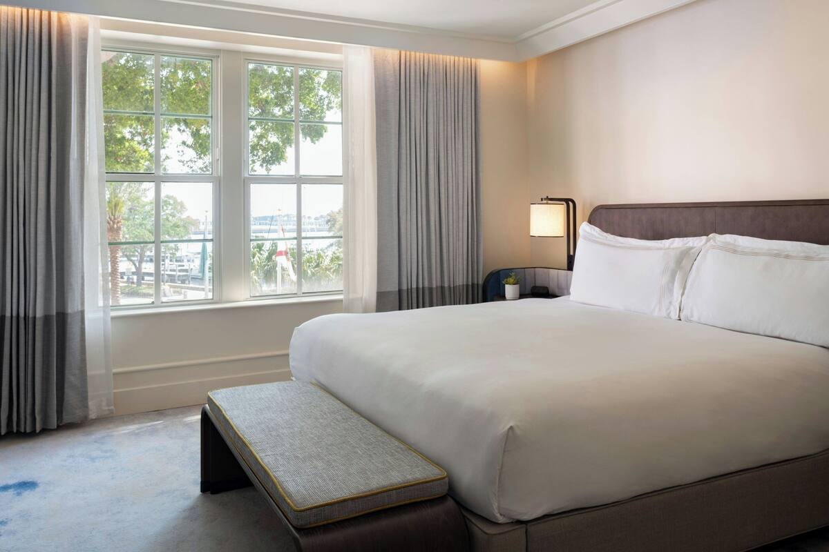 Ein Hotelzimmer mit einem großen Bett, einem Nachttisch, einer Lampe und einem Fenster mit Vorhängen.
