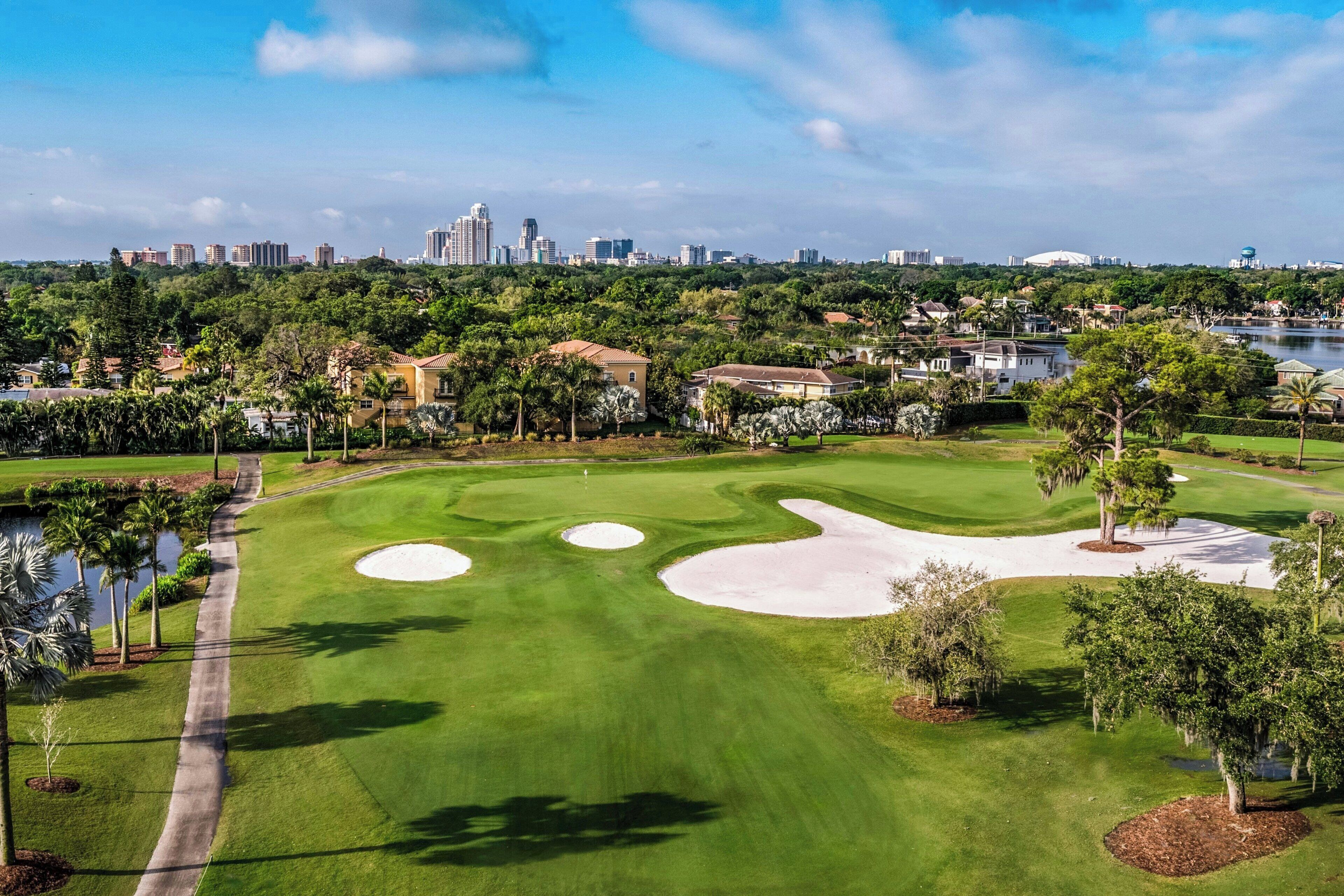 A golf course with a city skyline in the background, surrounded by greenery and water features.