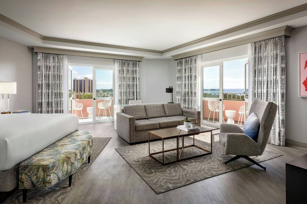 Ein modernes Hotelzimmer mit einem großen Bett, einer Couch, einem Couchtisch und einem Sessel. Das Zimmer verfügt über große Fenster mit Vorhängen, einen Blick auf die Stadt und einen Balkon mit Gartenmöbeln.