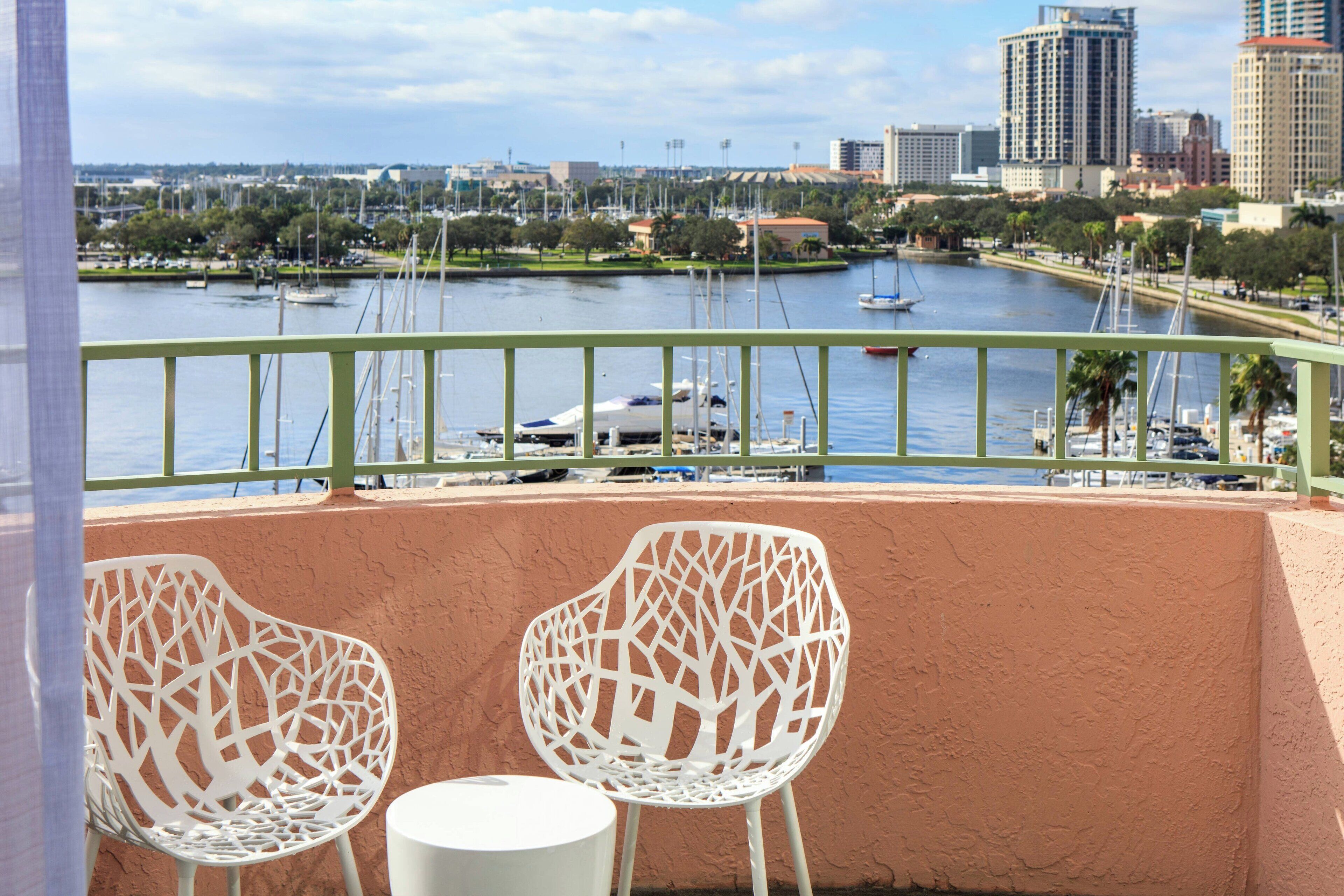 A balcony with two white chairs and a small table, overlooking a marina with boats and a cityscape in the background.