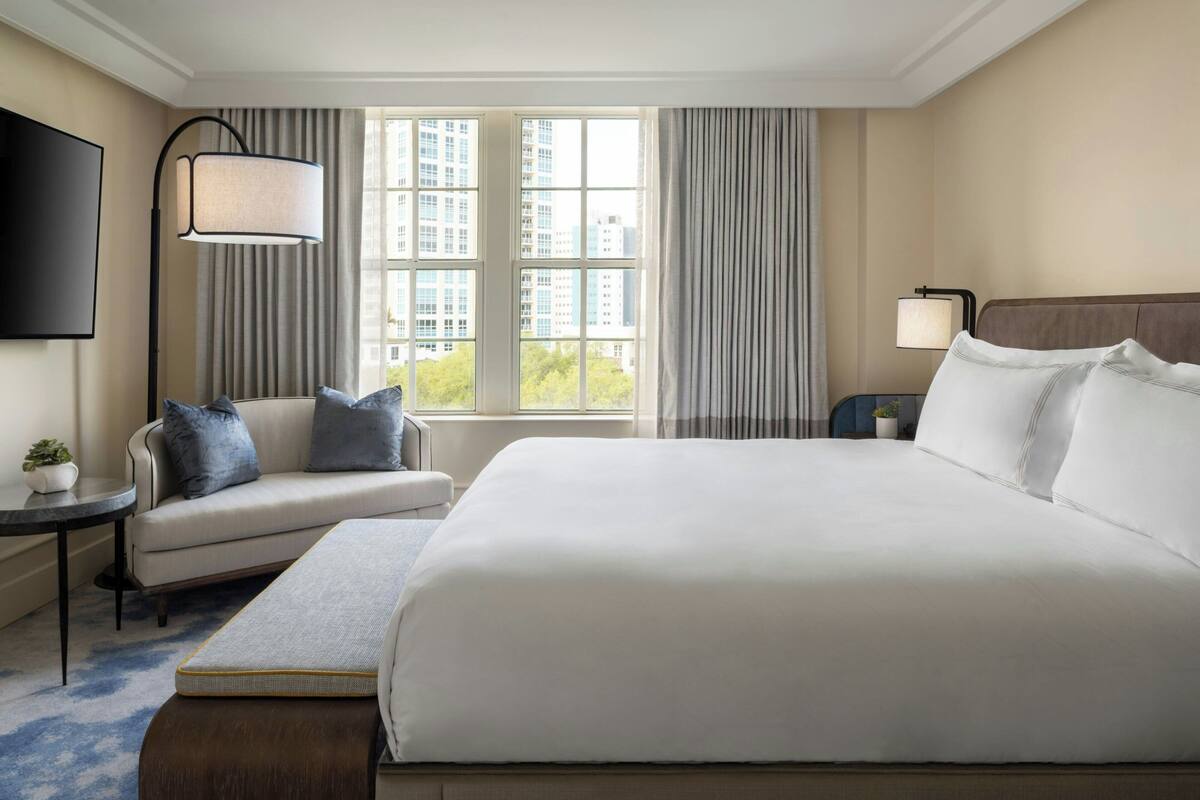 Ein Hotelzimmer mit einem großen Bett, einem Sofa, einem Fernseher, einer Lampe und Blick auf Gebäude durch das Fenster.