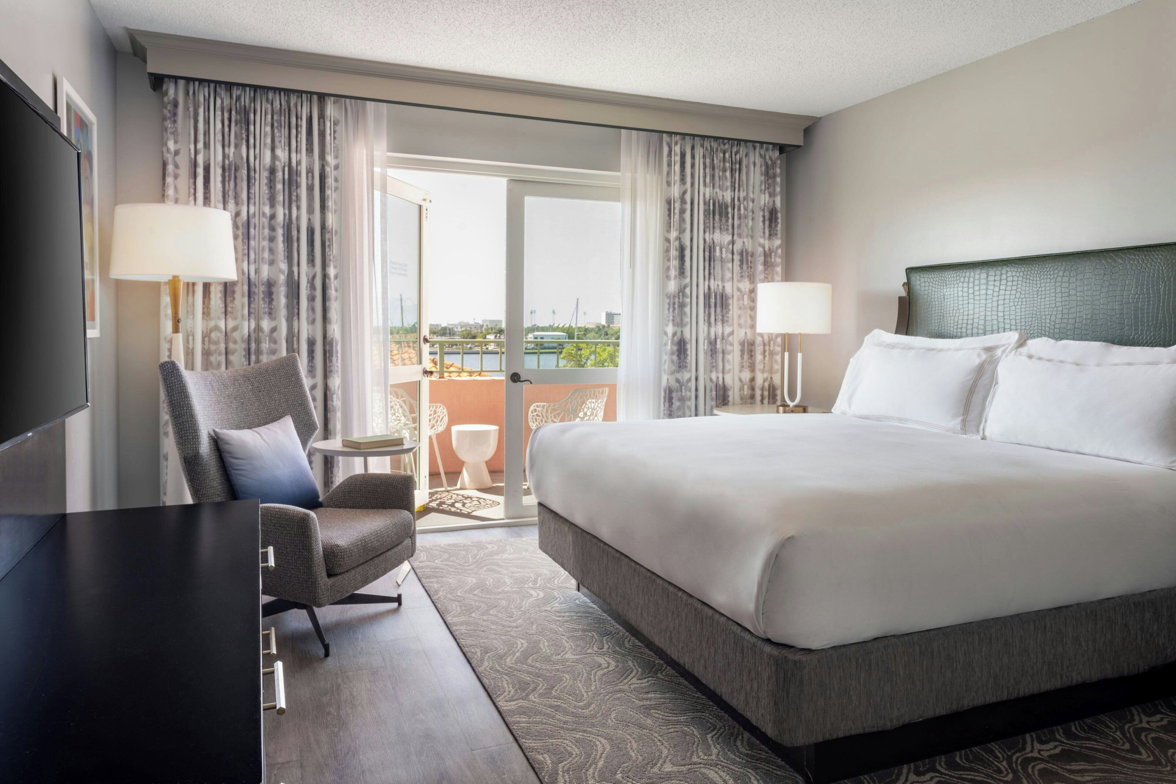 A hotel room with a large bed, a desk, a chair, two lamps, and a view of the outdoors through a glass door.