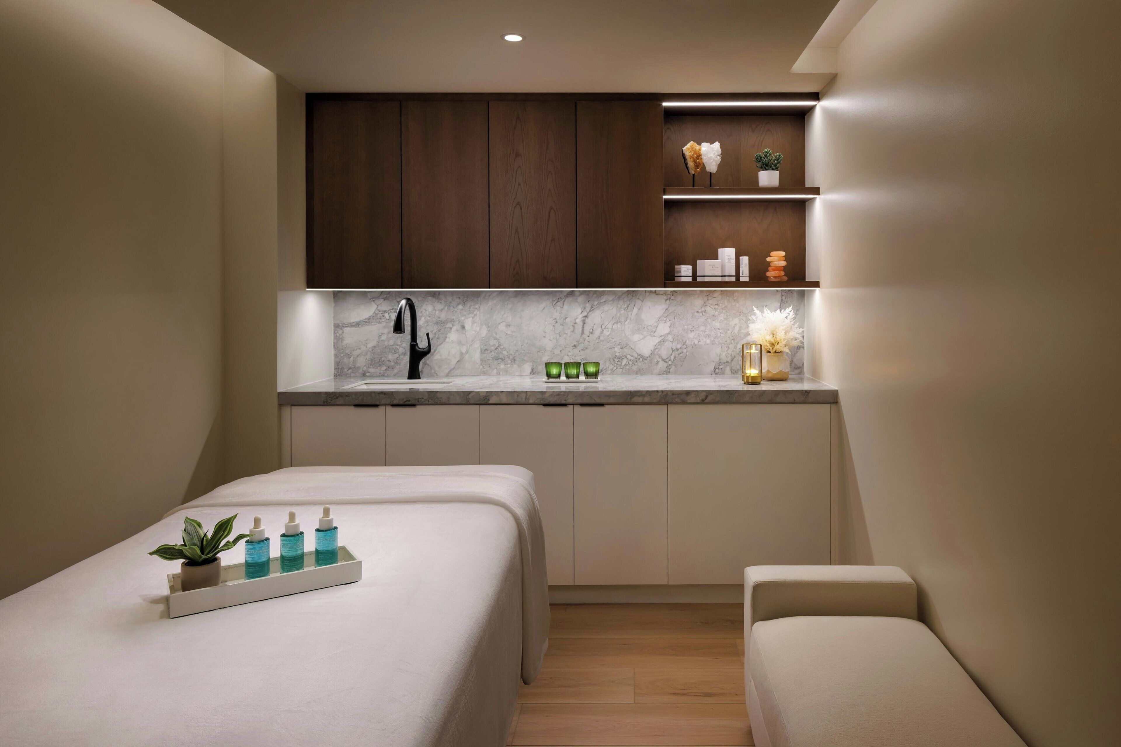 A compact room with a bed, a small seating area, a kitchenette with marble countertops, wooden cabinets, and a small shelf with decorative items.
