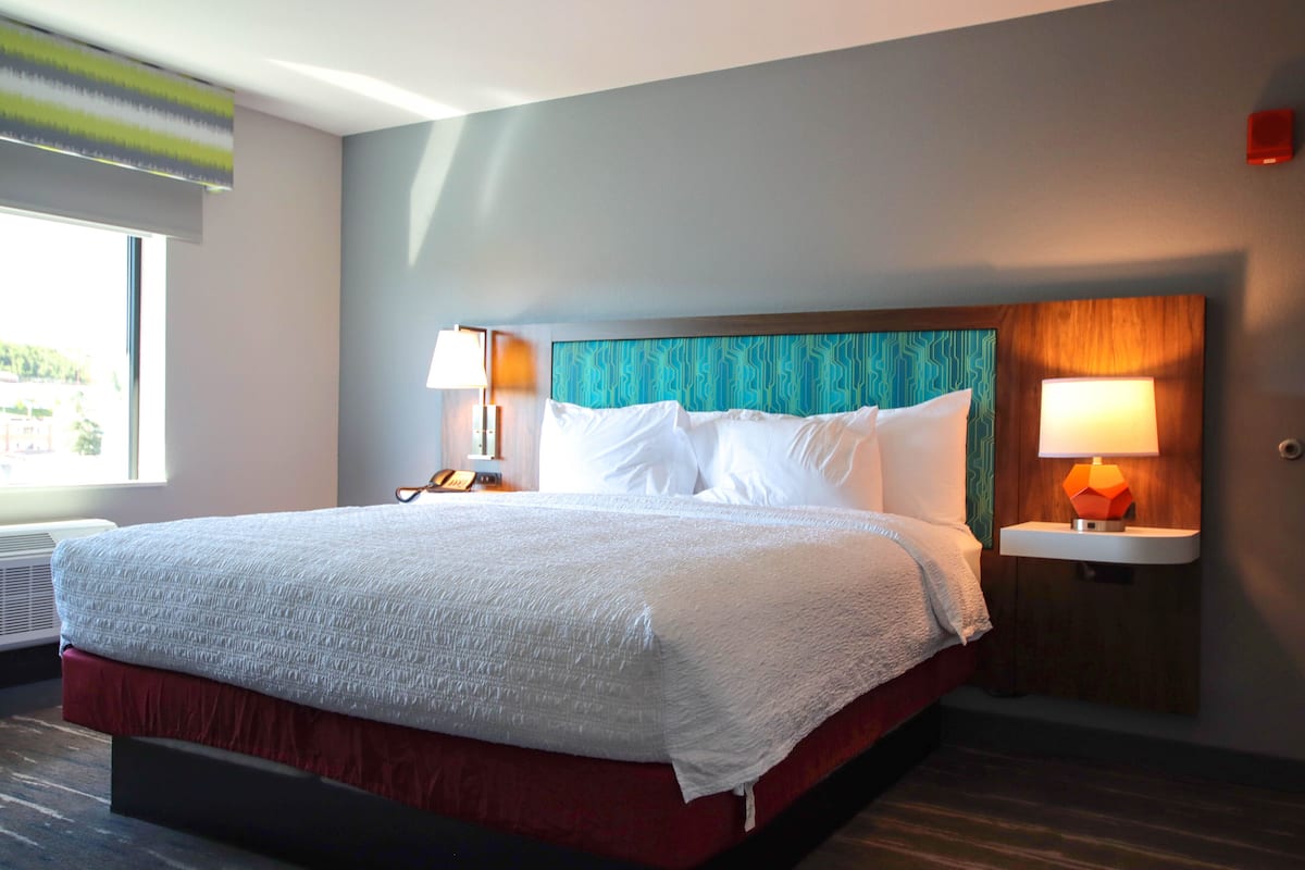 A hotel room with a large bed, two bedside tables with lamps, a window with blinds, and a view of greenery outside.