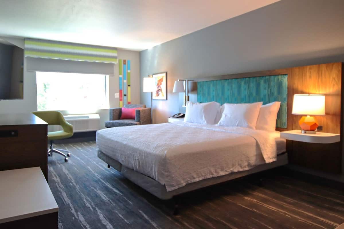 A hotel room with a large bed, a desk, a chair, a nightstand with a lamp, and a window with a view.
