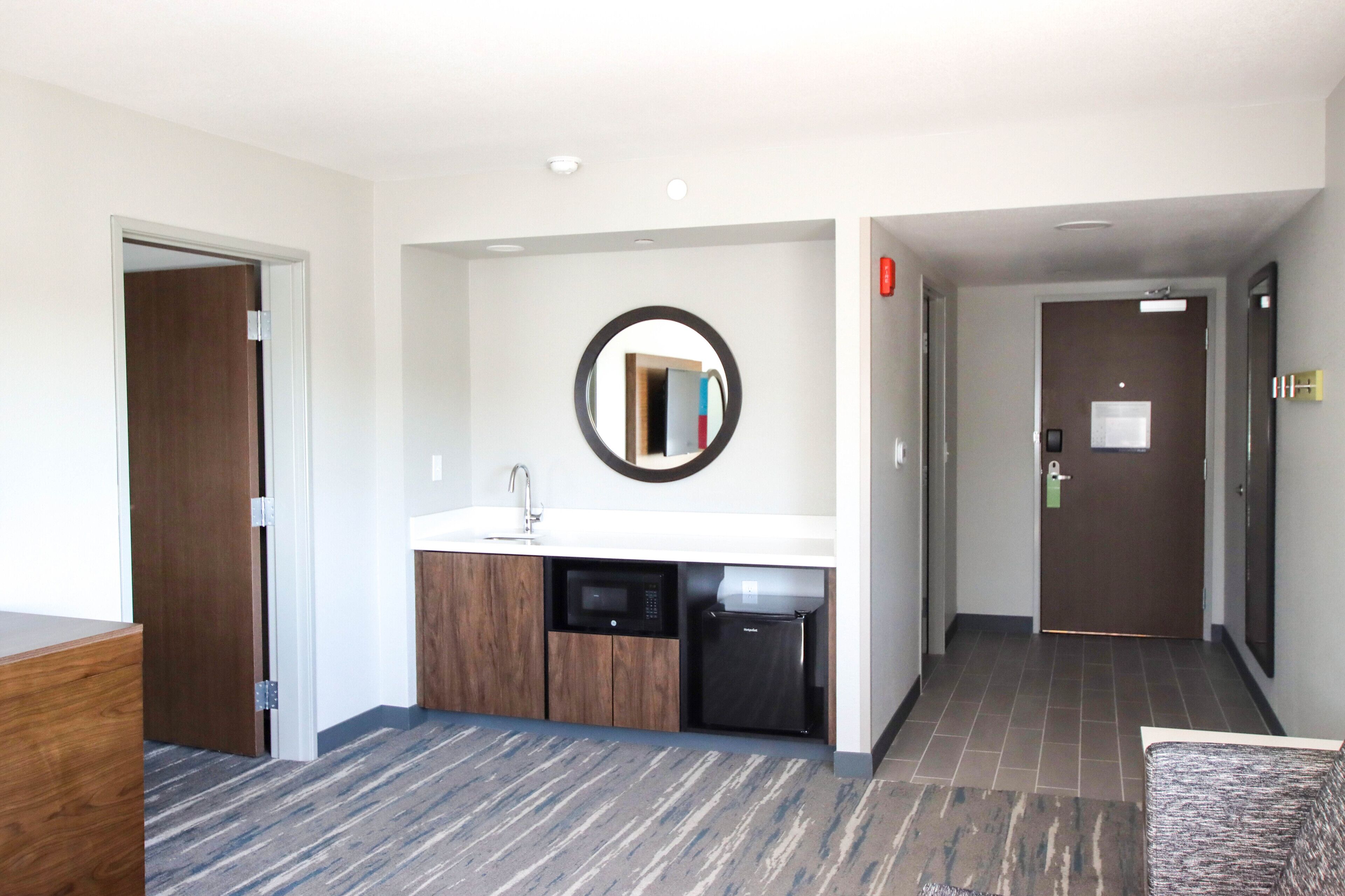 Una habitación de hotel moderna con un escritorio de madera, una cocina integrada con microondas y lavavajillas, un espejo redondo y una puerta con cerradura de llave electrónica.