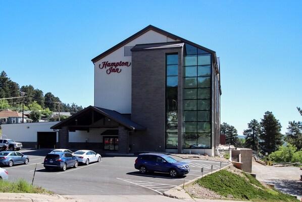 A Hampton Inn hotel with a modern design, featuring a large glass entrance and a parking lot with several cars.