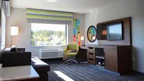 Una habitación de hotel moderna con sofá, escritorio, silla, televisión y ventana con vistas a edificios.