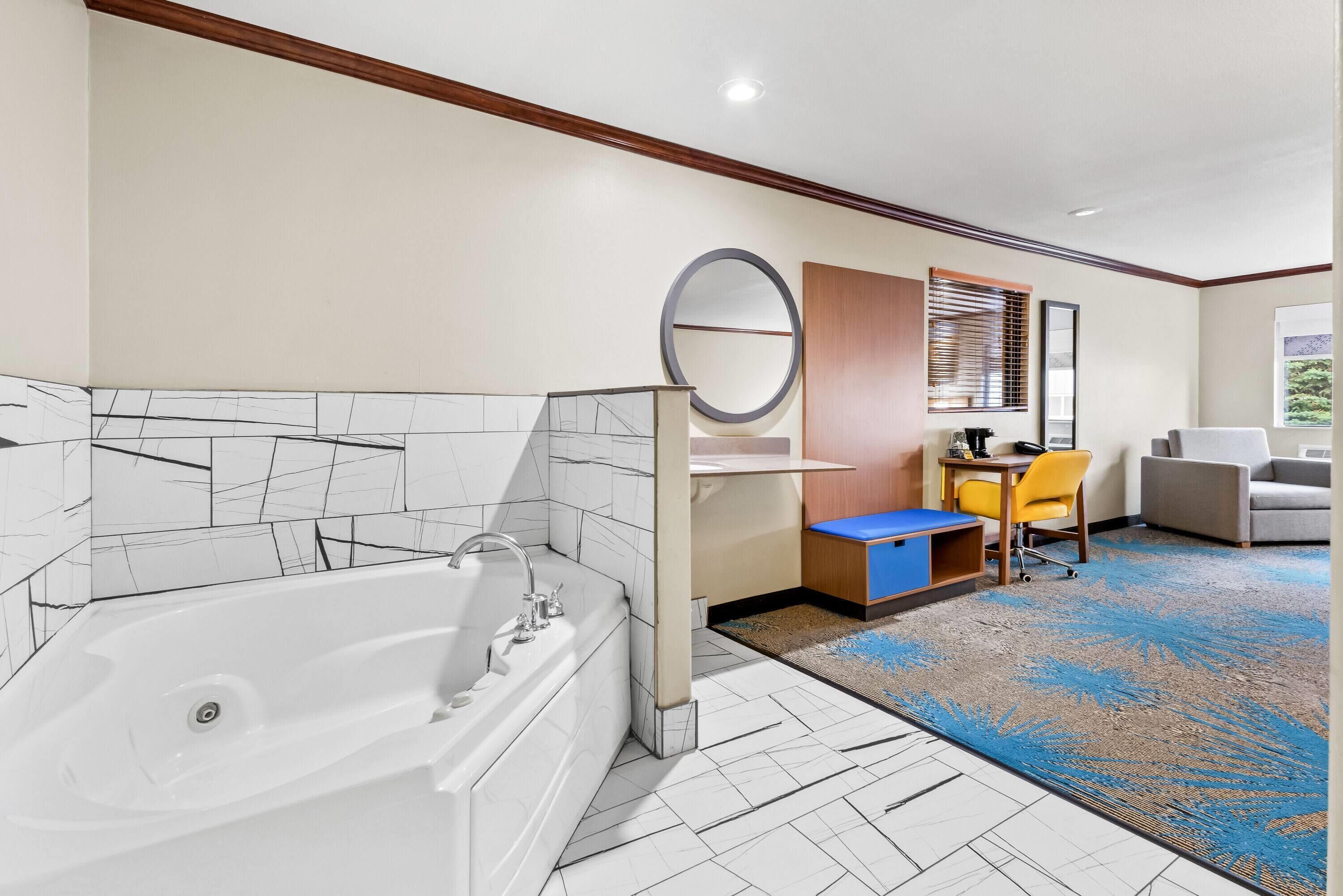 A modern bathroom with a bathtub, a sink, and a mirror. There is a desk with a chair and a window with blinds. The room has a carpeted floor and a view of greenery outside.