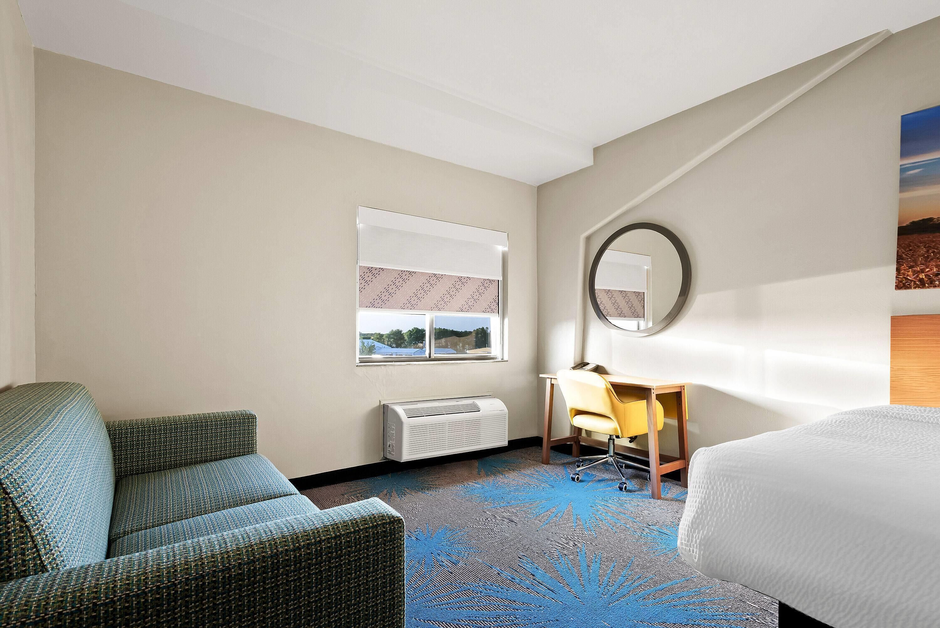 A hotel room with a bed, a sofa, a desk with a chair, a window with blinds, a wall-mounted mirror, and a carpet with a leaf pattern.