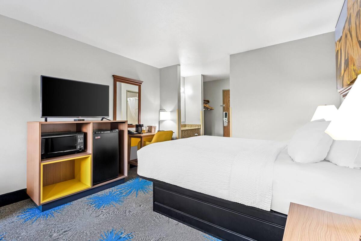 A hotel room with a large bed, a flat-screen TV, a microwave, a mini-fridge, a desk with a chair, and a bathroom visible through an open door.