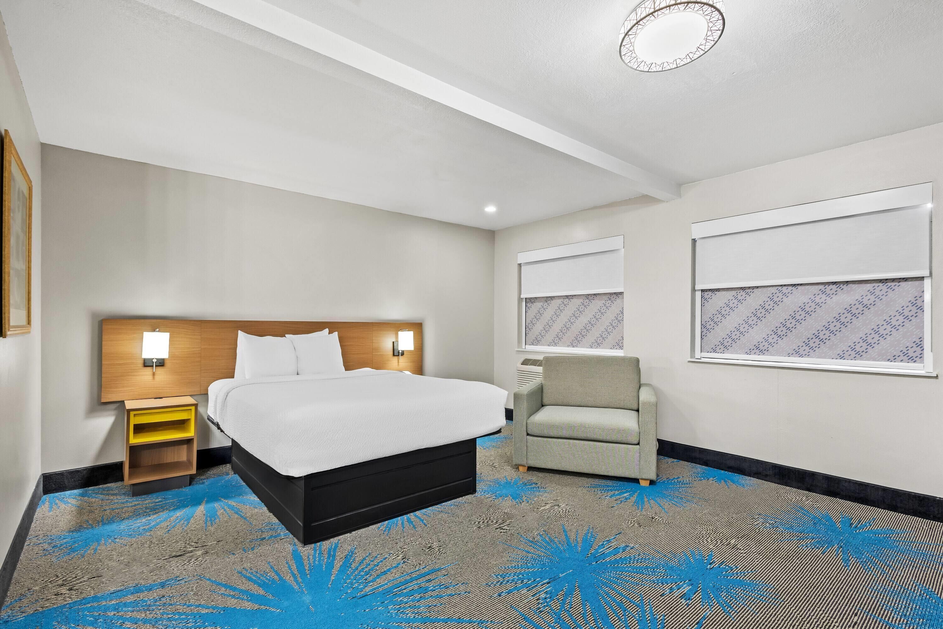 Una habitación de hotel con cama, una silla, una mesita de noche y una lámpara de pared.