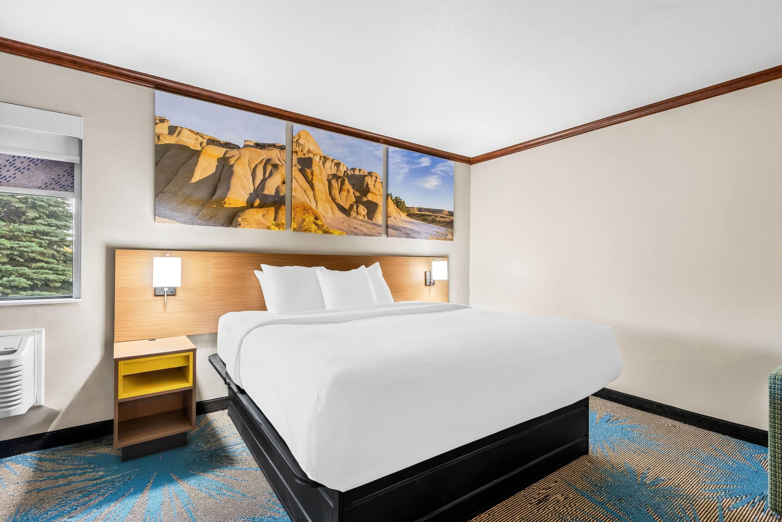 A hotel room with a bed, a nightstand, a wall-mounted lamp, and a large landscape painting above the bed.
