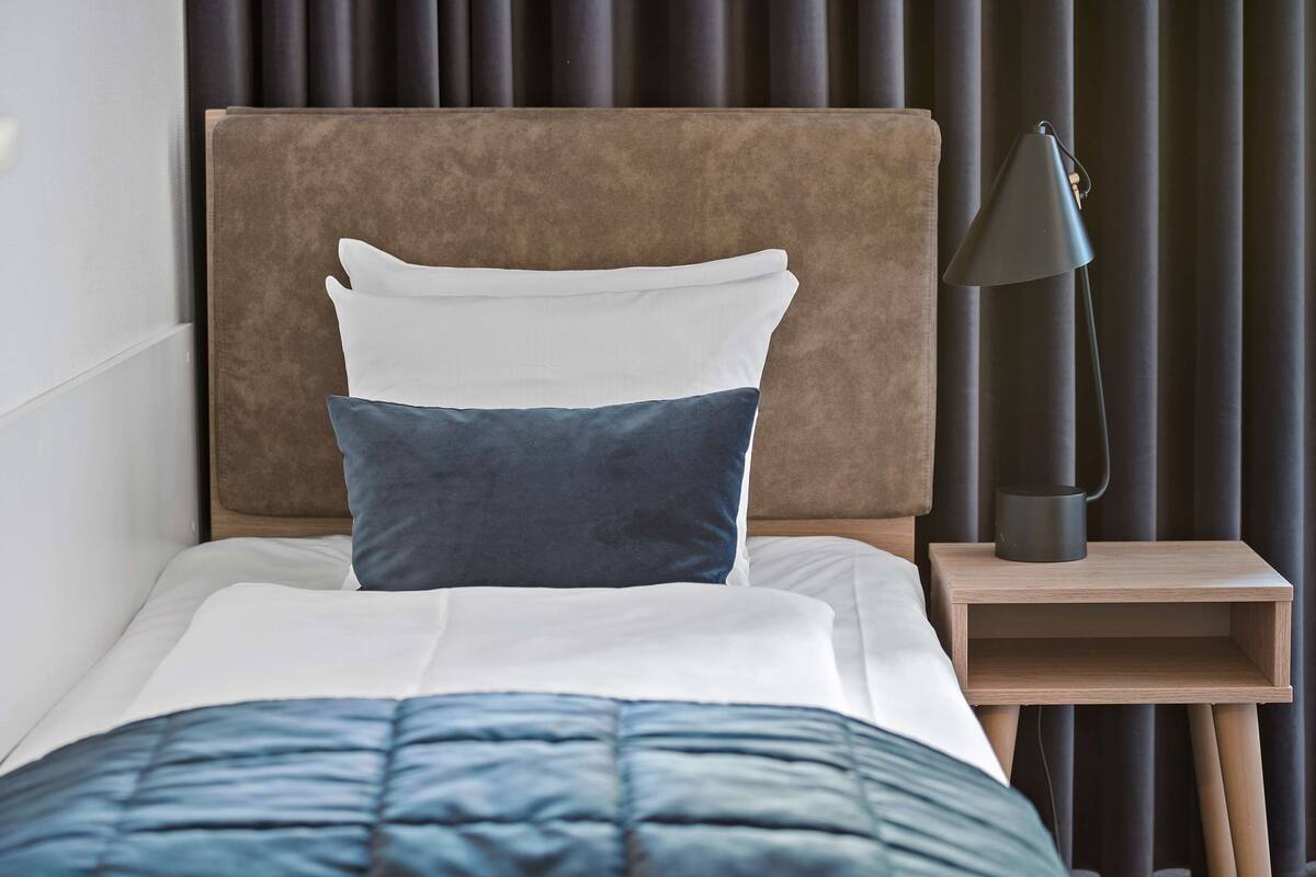 A neatly made bed with a blue quilt, a white pillow, and a dark blue pillow, accompanied by a black desk lamp on a wooden bedside table.