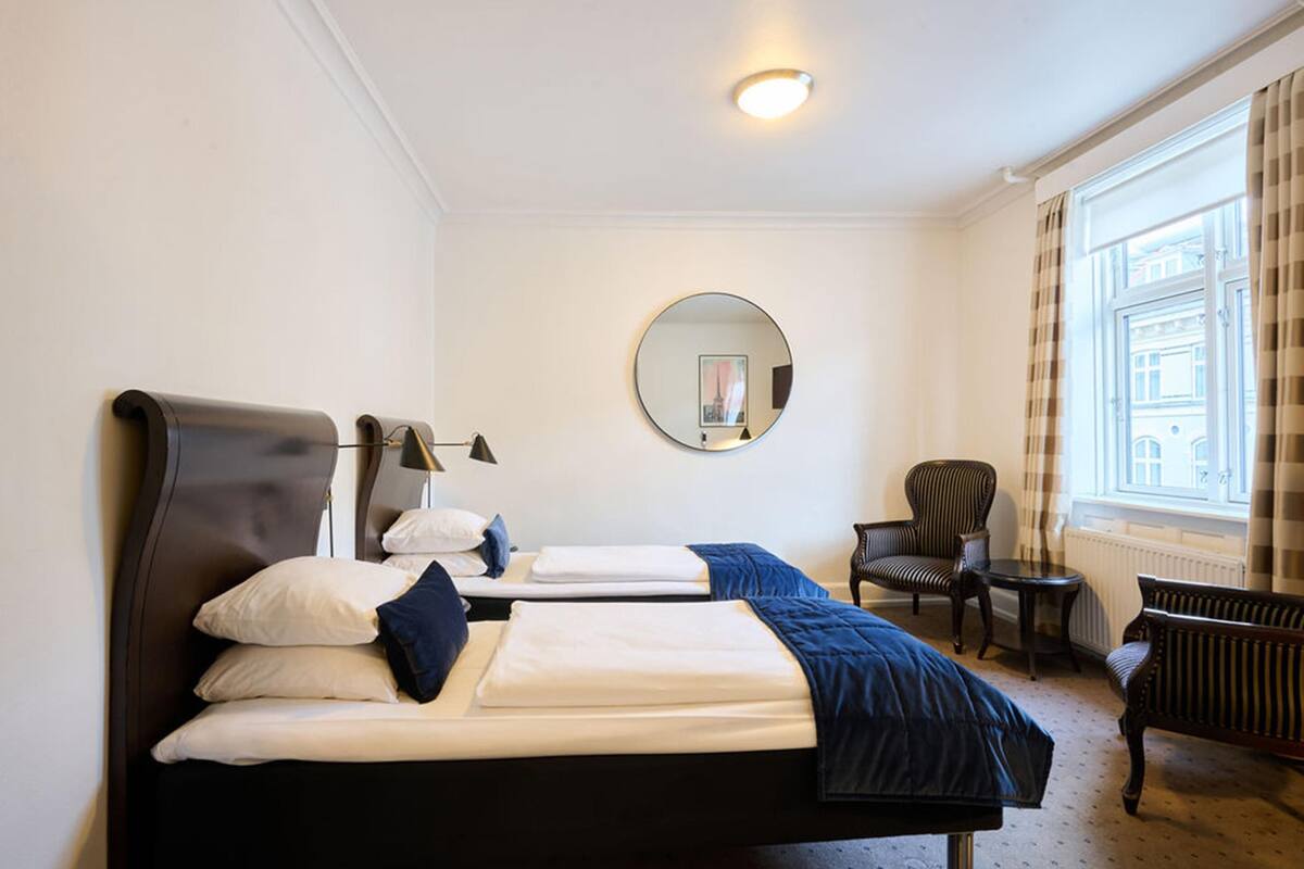 A hotel room with two beds, a round mirror, a chair, and a window with curtains.