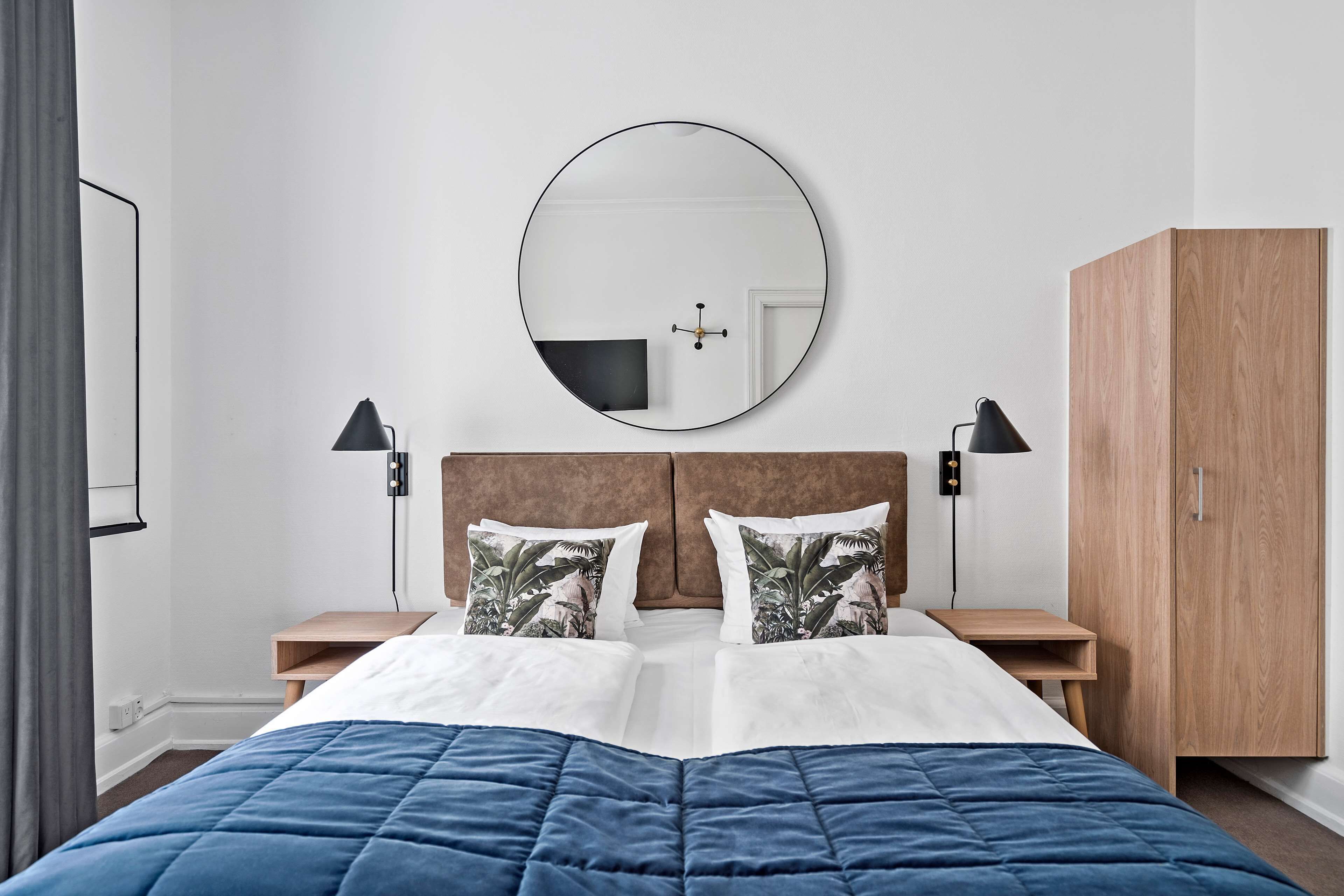 A neatly arranged bedroom with a large bed, a round mirror, bedside lamps, and a wooden wardrobe.