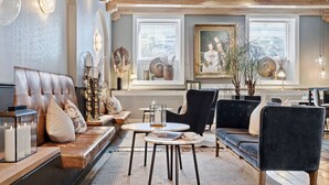 A modern living room with a leather sofa, a round table, and a black armchair. There are two windows with blinds, a chandelier, and decorative vases. - Best Western Plus Hotel Hebron (Copenhagen)
