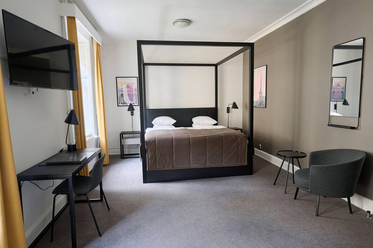 A modern bedroom with a four-poster bed, a desk with a chair, a small round table, and a chair.
