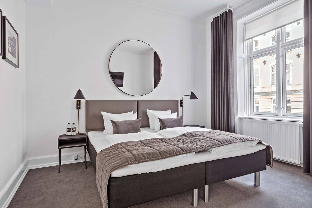 A modern hotel room with a large bed, a round mirror, bedside lamps, and a view of the city through the window.