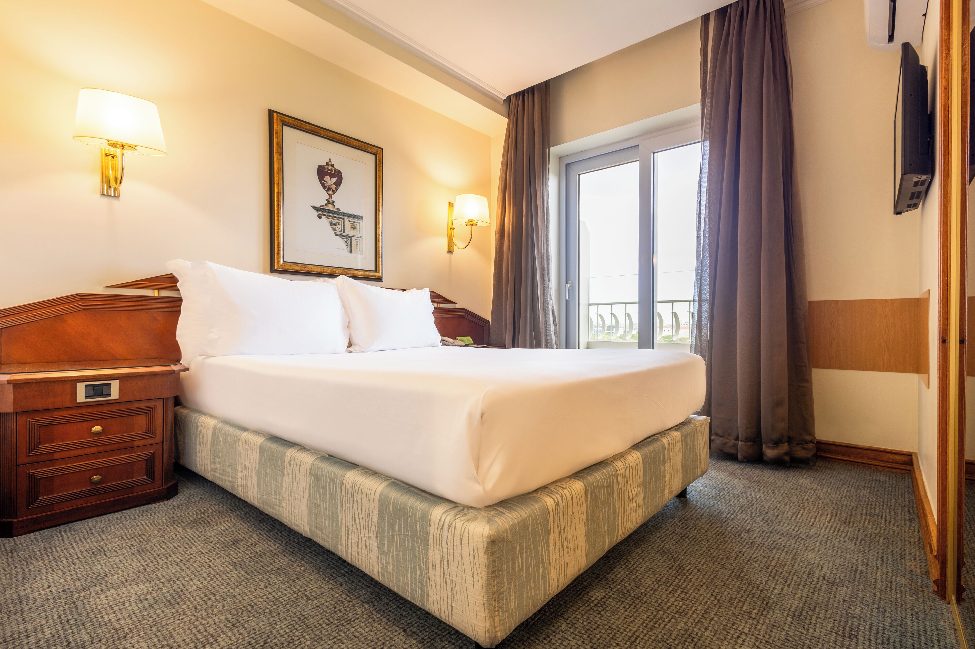 A hotel room with a large bed, two bedside tables, a framed picture on the wall, a window with curtains, and a flat-screen TV mounted on the wall.