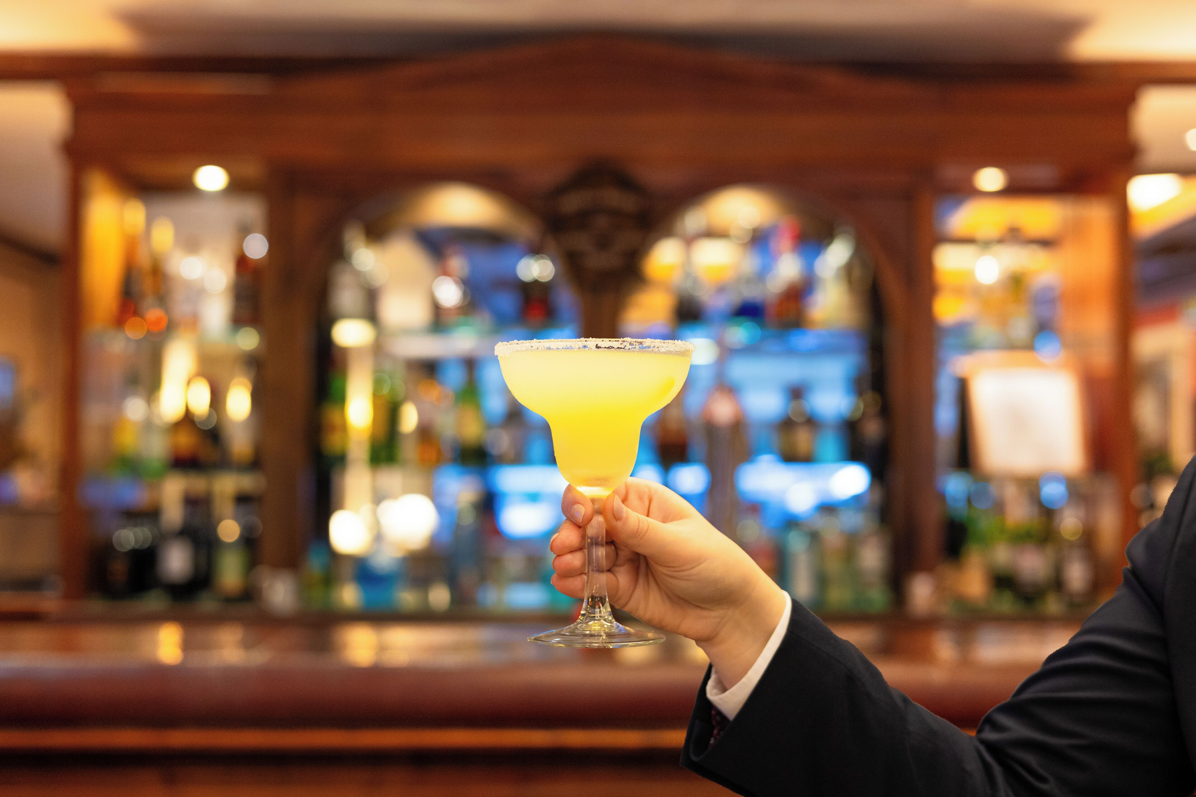 A hand holding a margarita glass with a salted rim, set against a blurred bar background with bottles and shelves.