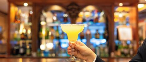 A hand holding a margarita glass with a salted rim, set against a blurred bar background with bottles and shelves.