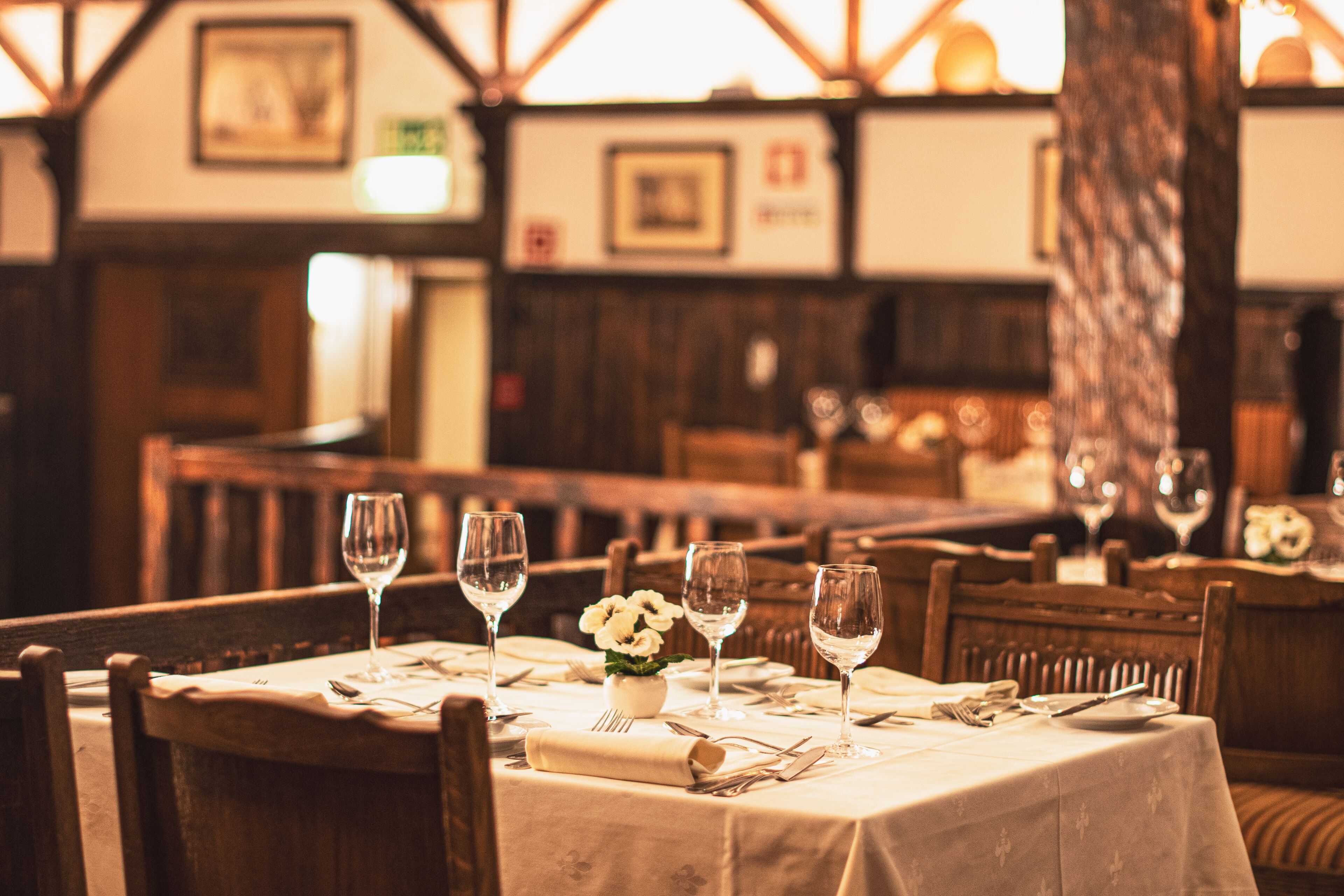 A dining area with wooden tables set for a meal, featuring white tablecloths and wine glasses.