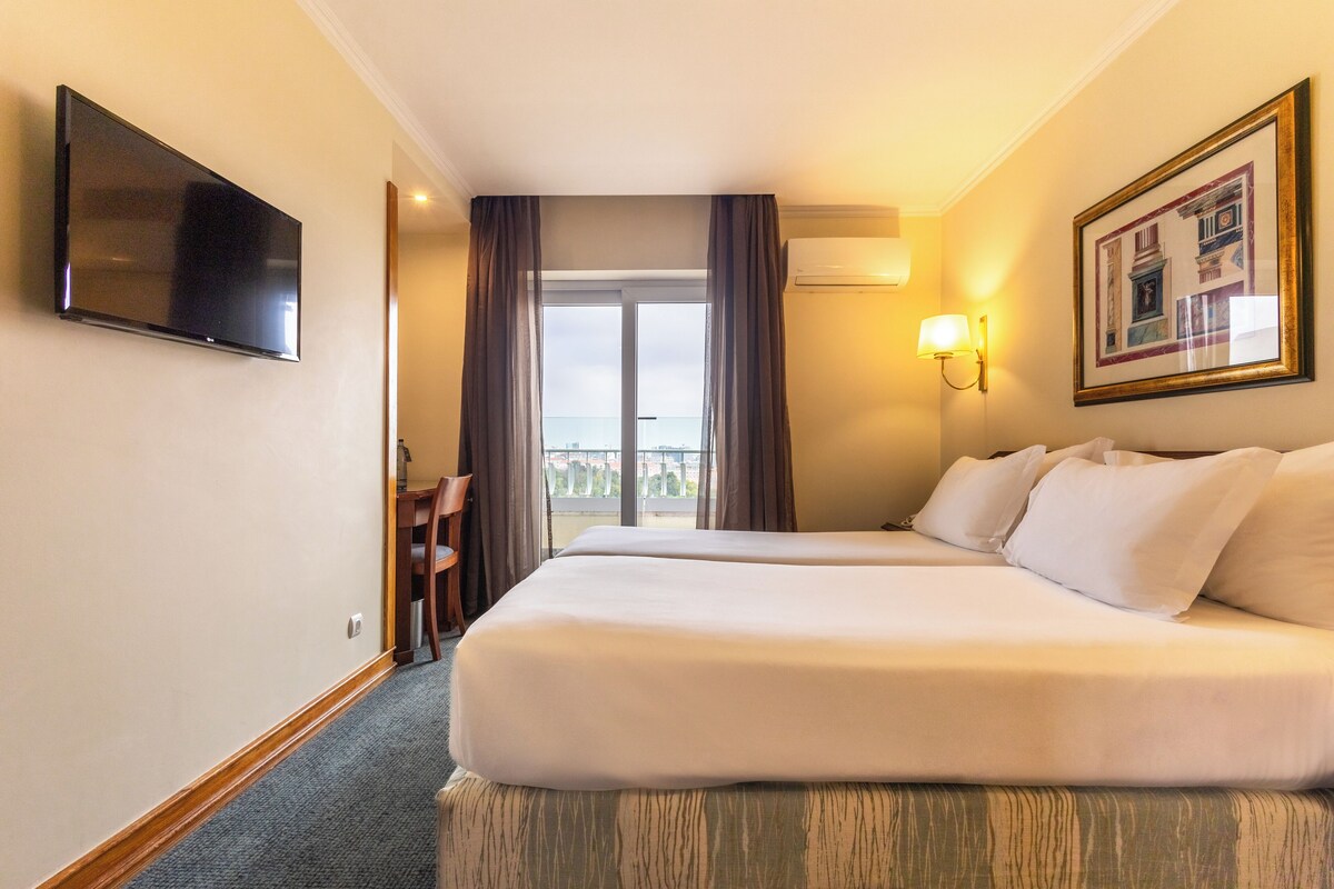 A hotel room with two beds, a TV, a desk, and a balcony view.