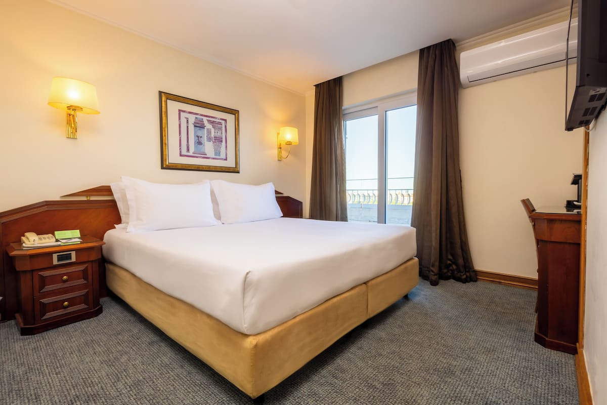 A hotel room with a large bed, a nightstand with a telephone, a framed picture on the wall, and a view of the outdoors through a window with curtains.