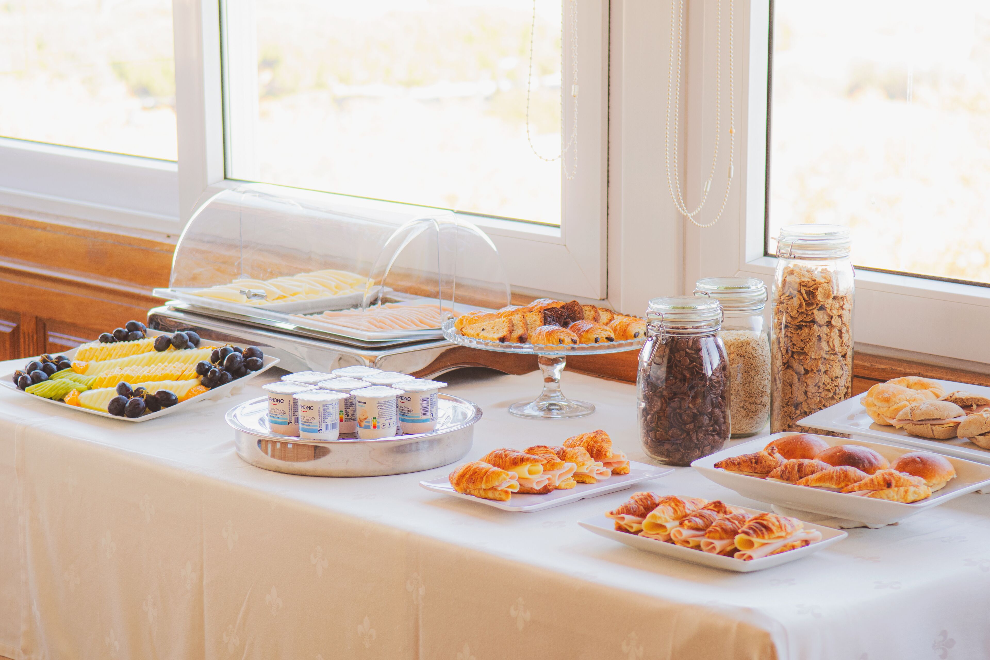 Mesa de buffet com diversos alimentos e bebidas, com vista para árvores através de uma janela.