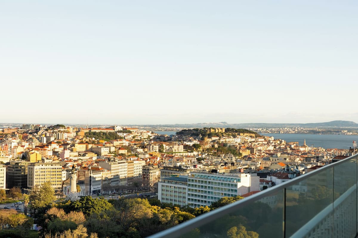 A panoramic view of a coastal city with historical architecture, a prominent hill, and a body of water in the distance.