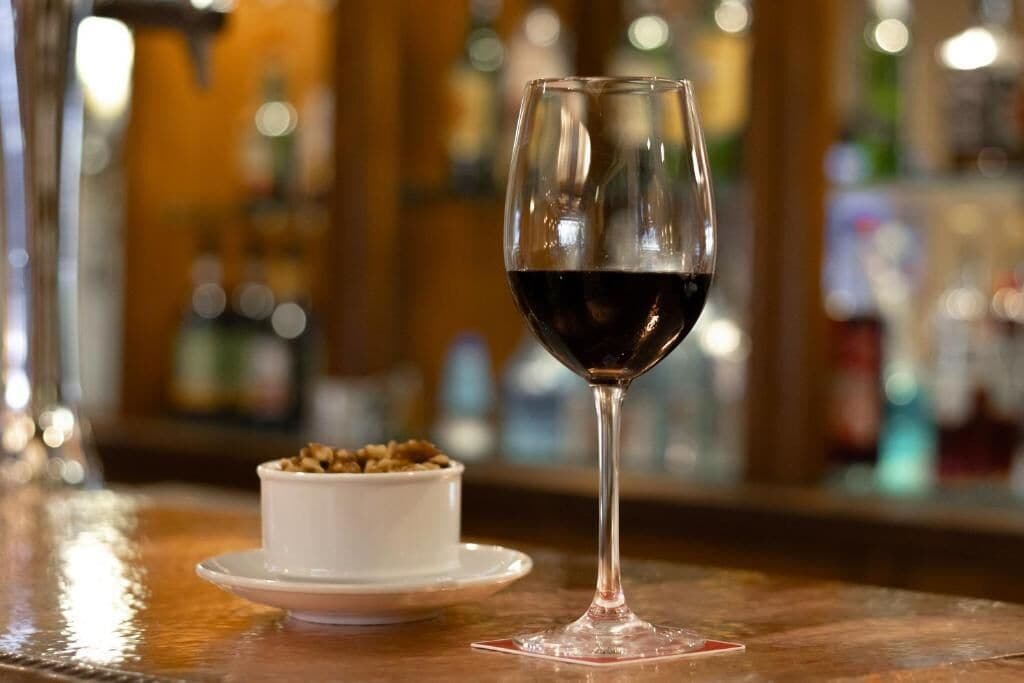 A glass of red wine on a bar counter with a small white dish of nuts.
