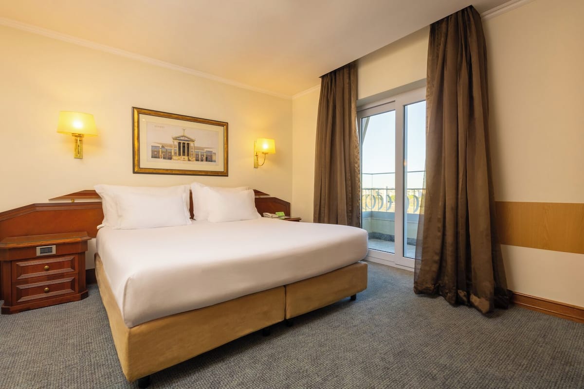 A hotel room with a large bed, two bedside lamps, a nightstand, a framed picture on the wall, and a view of the outdoors through a sliding glass door.