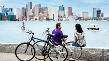 A waterfront view with a city skyline in the background, a boat on the water, and bicycles parked by the edge.