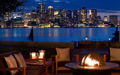 A waterfront patio with outdoor seating, a fire pit, and a city skyline in the background.