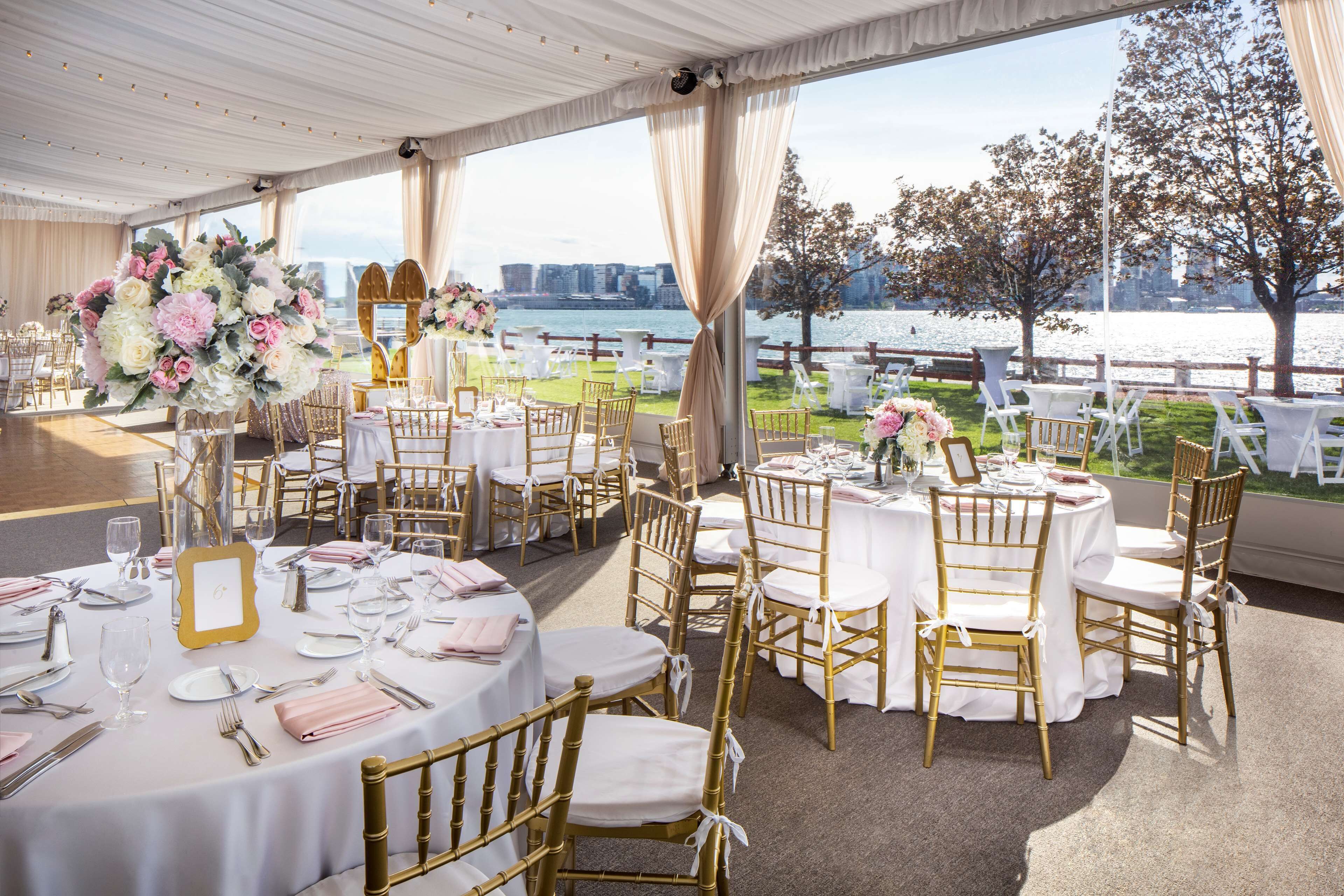 An outdoor wedding venue with round tables, gold chairs, and floral centerpieces, overlooking a body of water and city skyline.
