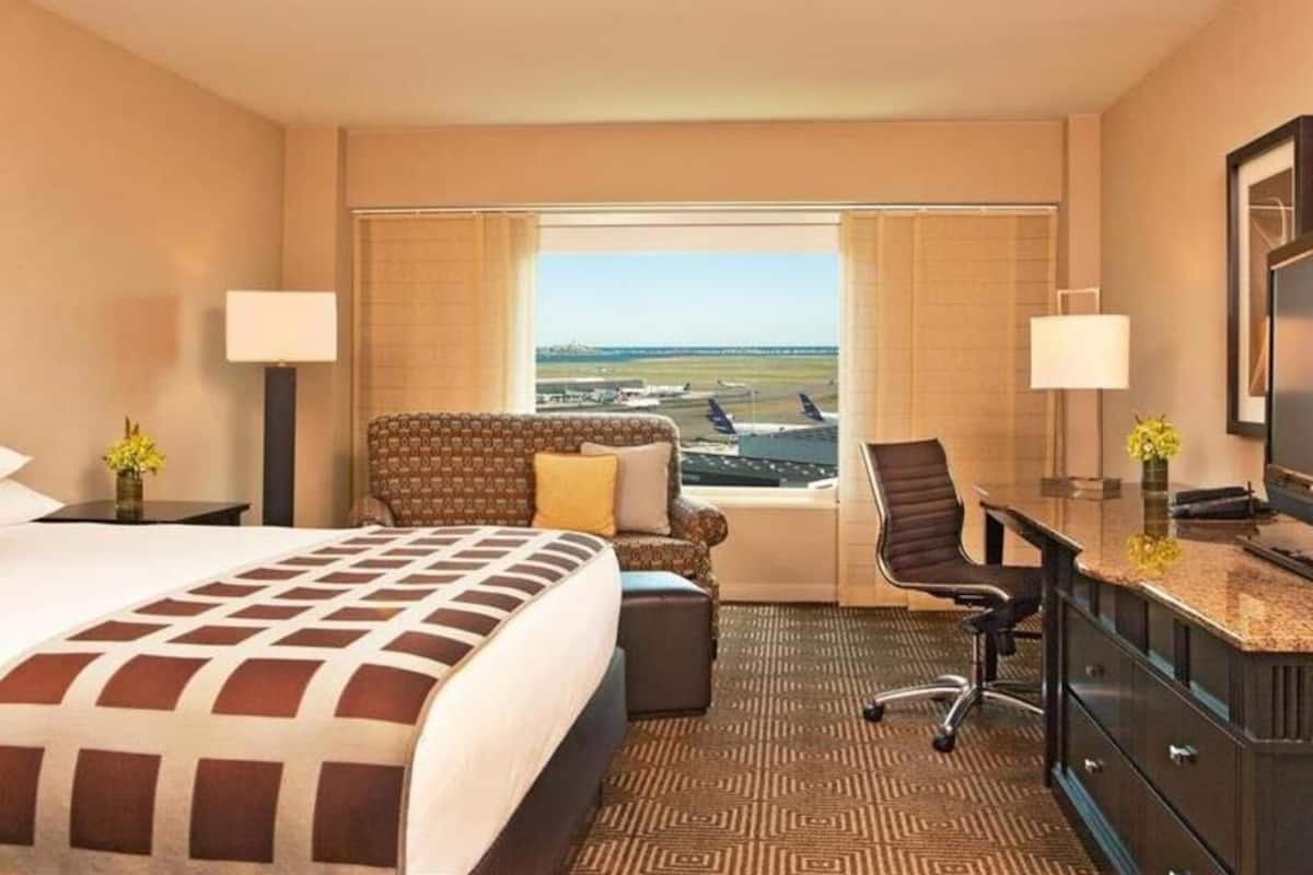 A hotel room with a bed, a desk, a chair, and a view of an airport.