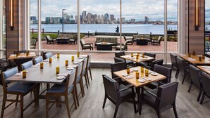 Een restaurant met tafels en stoelen, uitzicht op een stadshemelbeeld over een wateroppervlak en een terras.