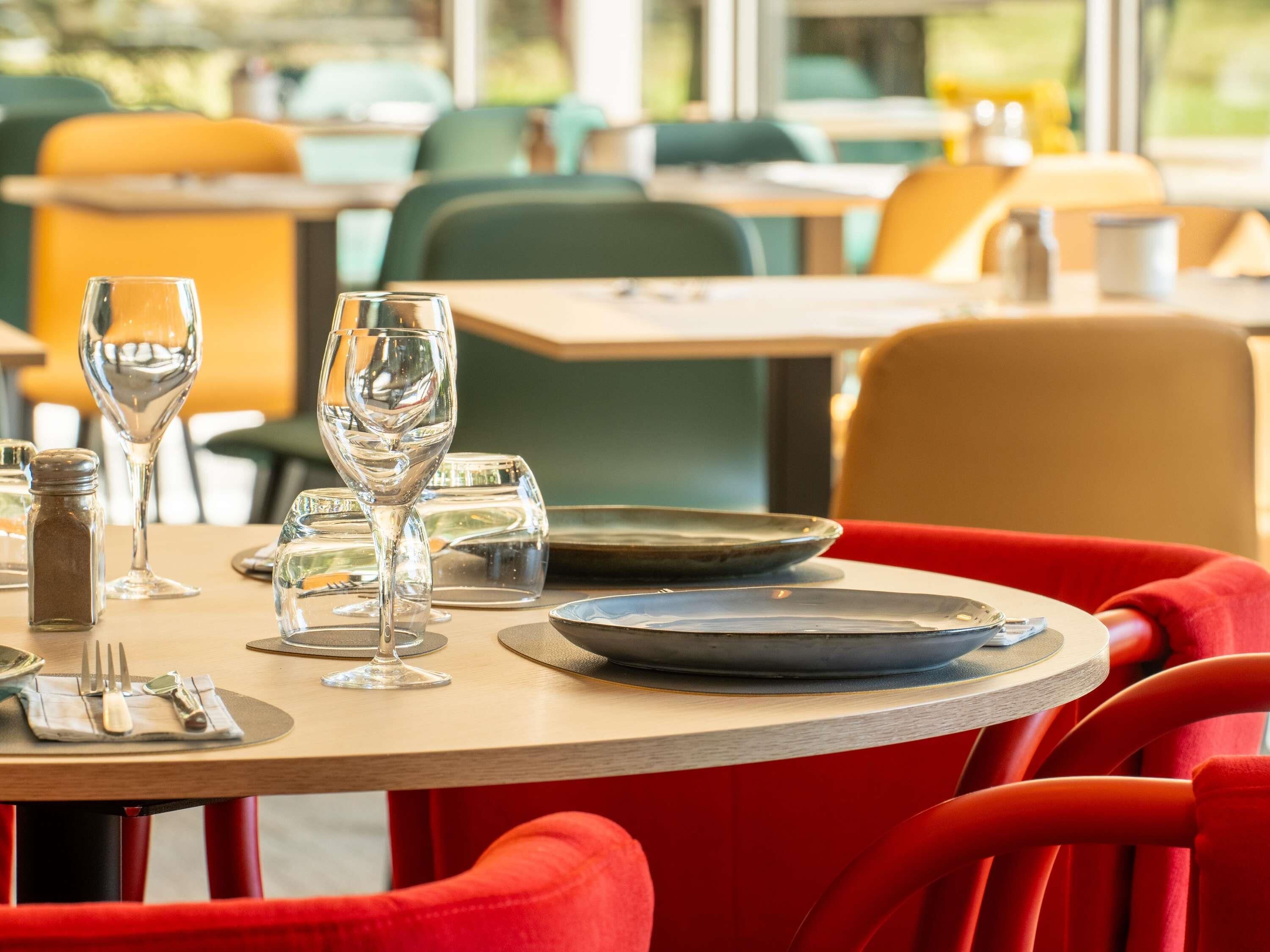 A dining table set with plates, glasses, and cutlery, surrounded by red upholstered chairs in a restaurant with a modern interior.