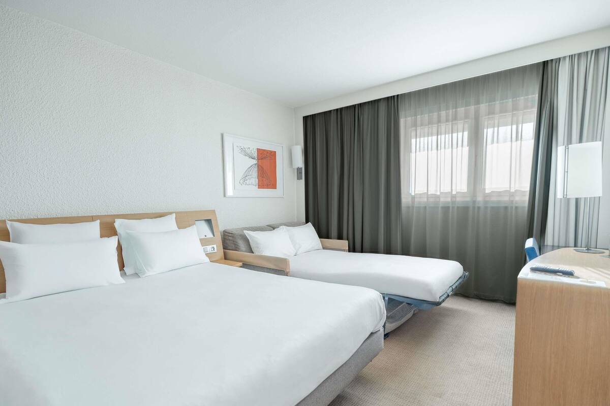 Quarto de hotel com duas camas, uma escrivaninha, uma luminária e uma janela com cortinas.