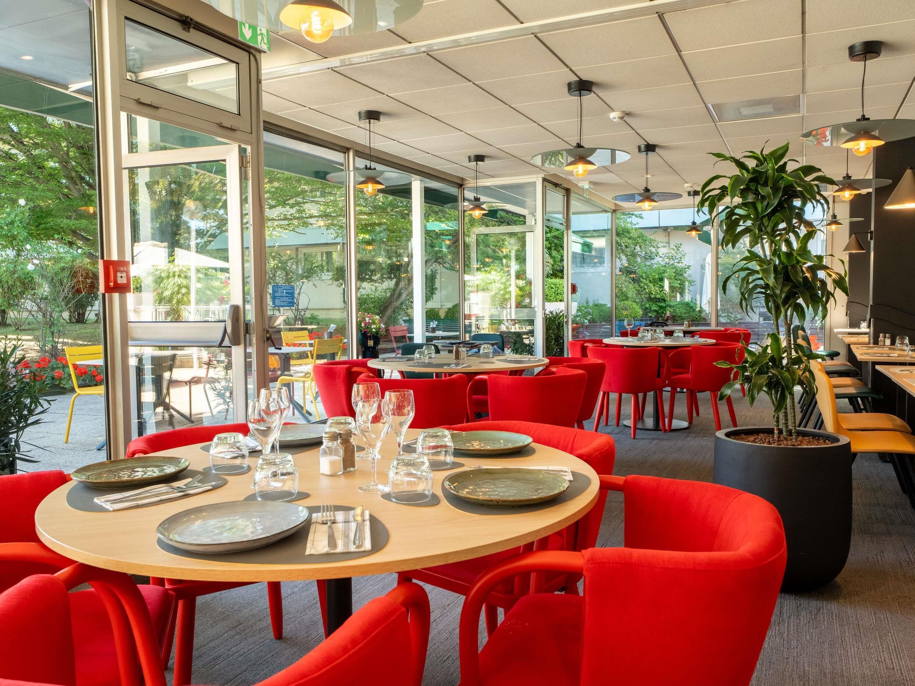A restaurant with round tables, red chairs, and large windows offering a view of the outdoors.