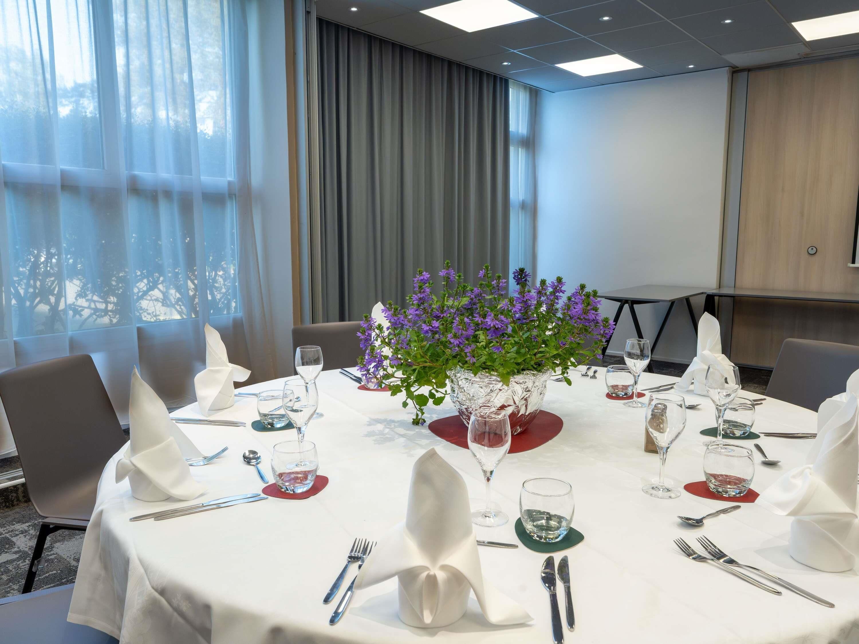 Ein Konferenzraum mit rundem Tisch, gedeckt für eine Besprechung, einer floralen Tischdekoration und einer Projektionsleinwand im Hintergrund.