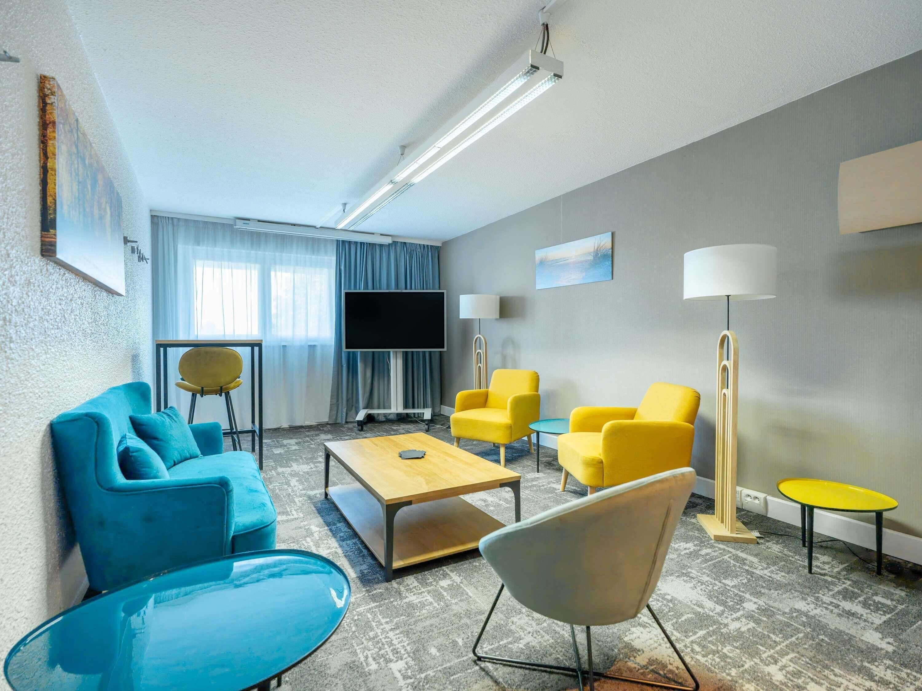 A modern hotel room with a sofa, chairs, a coffee table, a TV, and a lamp.