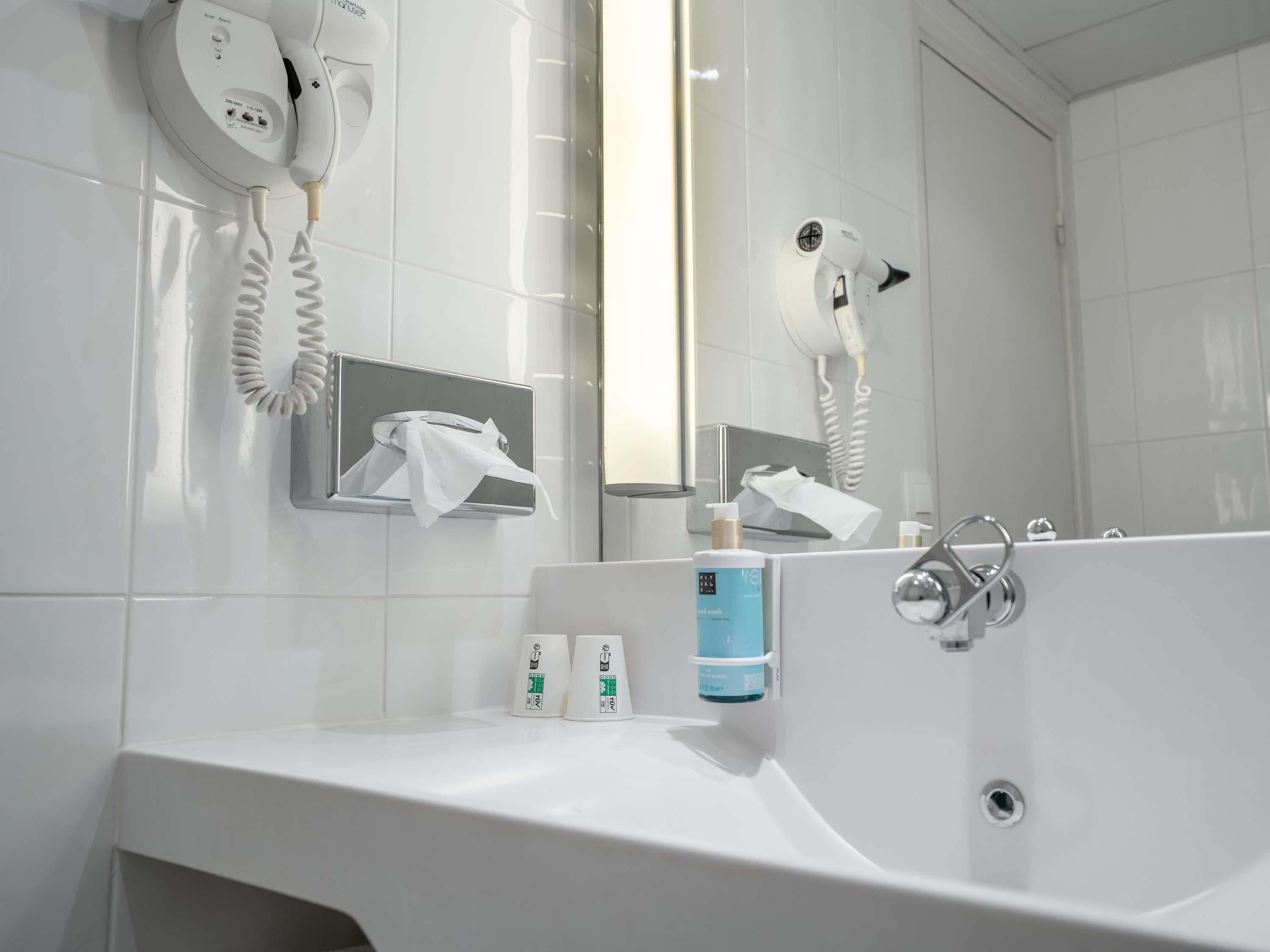 Une salle de bain moderne avec un grand lavabo, deux sèche-cheveux fixés au mur, un miroir et un distributeur de serviettes.