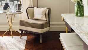 Una habitación con vistas a edificios, un cuadro en la pared, una silla, una mesa con un jarrón de flores y una lámpara.