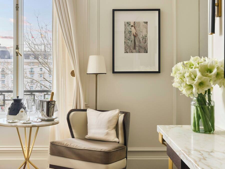 A room with a view of buildings, a framed picture on the wall, a chair, a table with a vase of flowers, and a lamp.