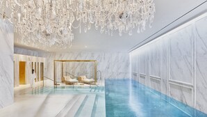 Un área de piscina interior lujosa con paredes de mármol, una gran lámpara de araña y un ambiente tipo spa con sillones reclinables y una bañera de hidromasaje con cerramiento de vidrio.