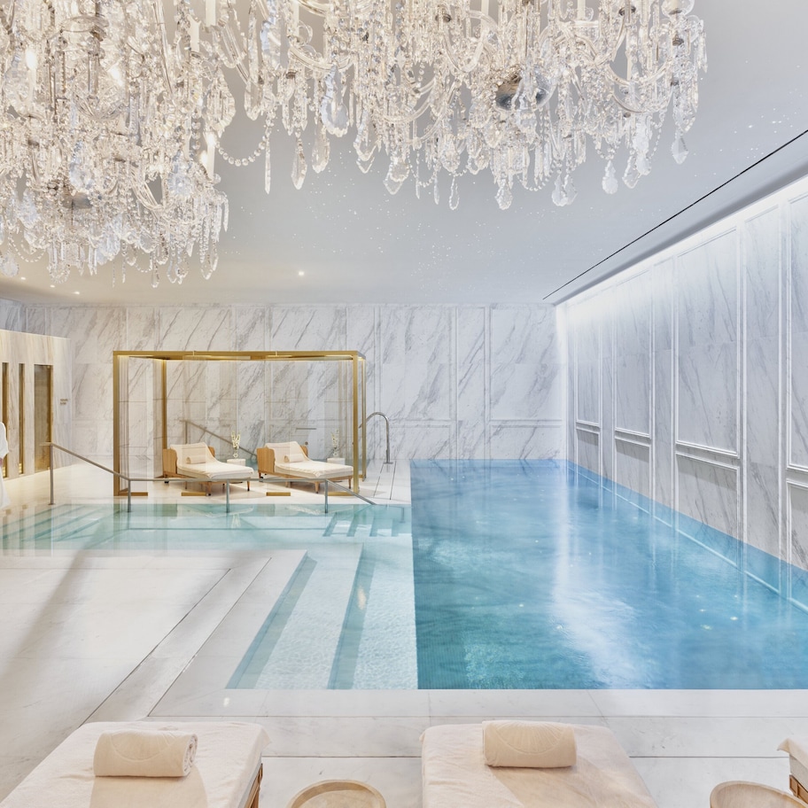 A luxurious indoor pool area with marble walls, a large chandelier, and a spa-like setting with loungers and a glass-enclosed hot tub.