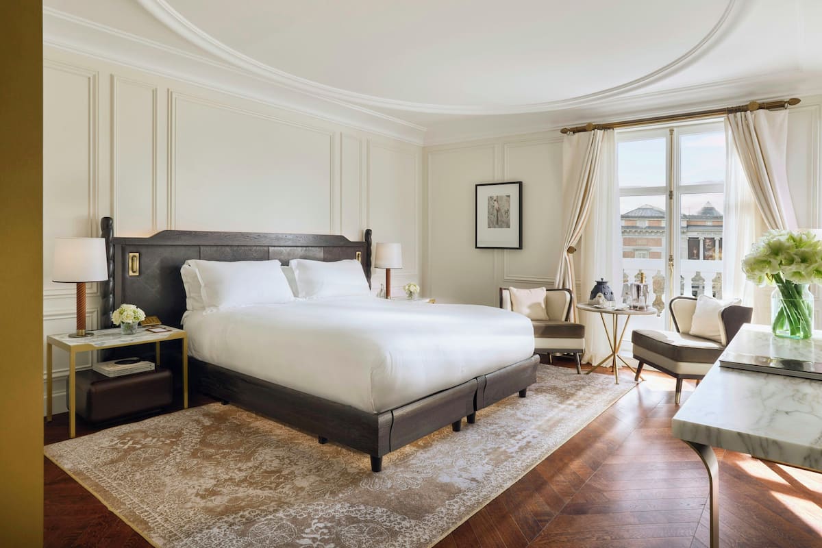 A hotel room with a large bed, a desk, a chair, a small table, and a window with curtains.