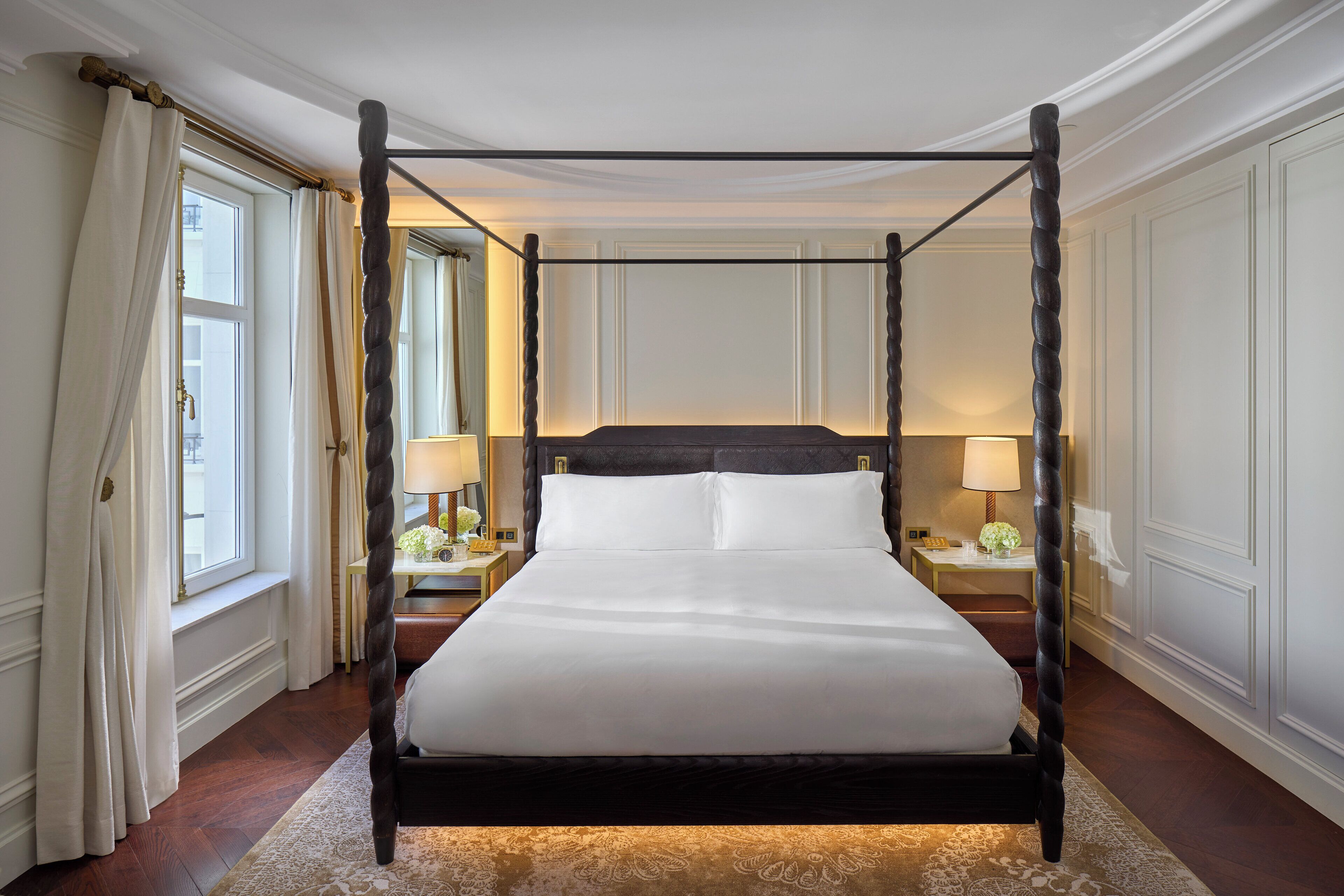 Una cama con dosel cubierta con sábanas blancas en una habitación con piso de madera, paredes blancas y dos mesitas de noche con lámparas.